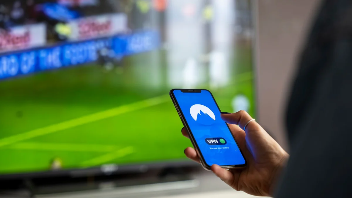 A person holding a smartphone with a VPN app, streaming sports on TV, illustrating iptv smarters pro android